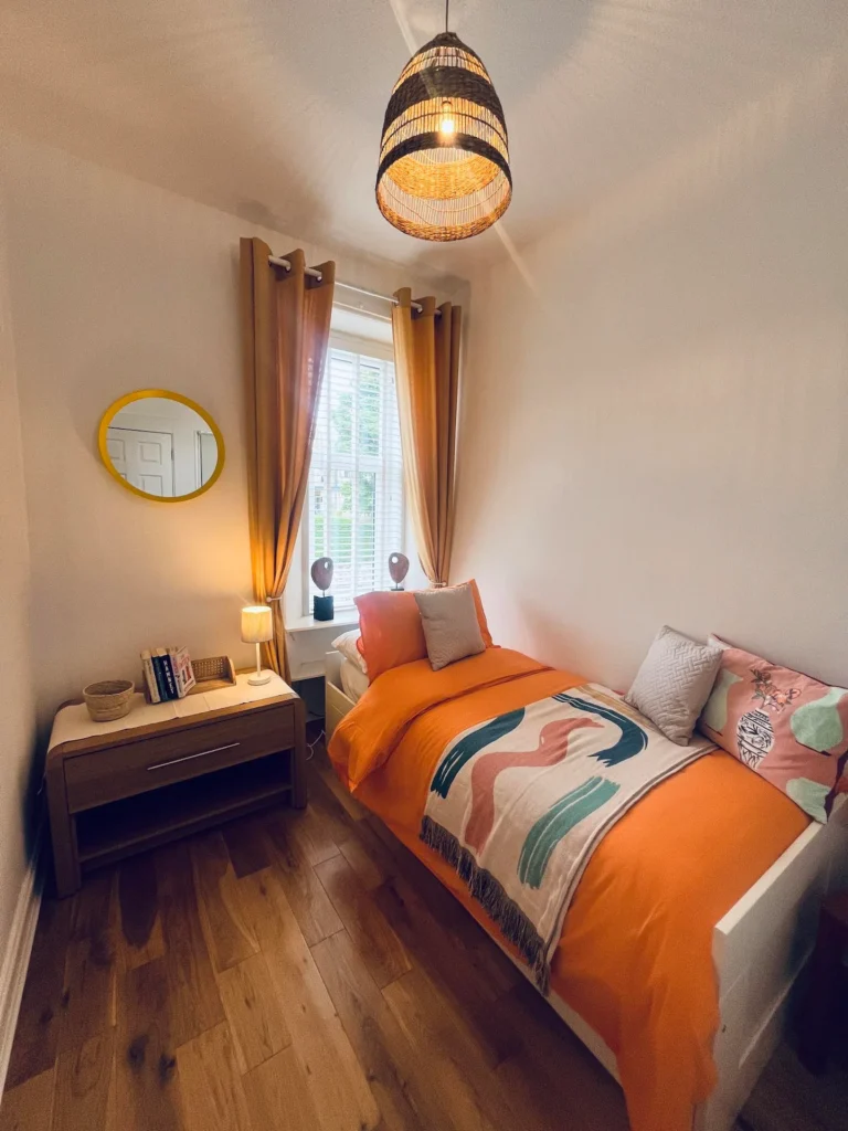 Cozy bedroom with orange bedding, wooden floor, wicker lamp, and soft lighting, creating a warm and inviting atmosphere.