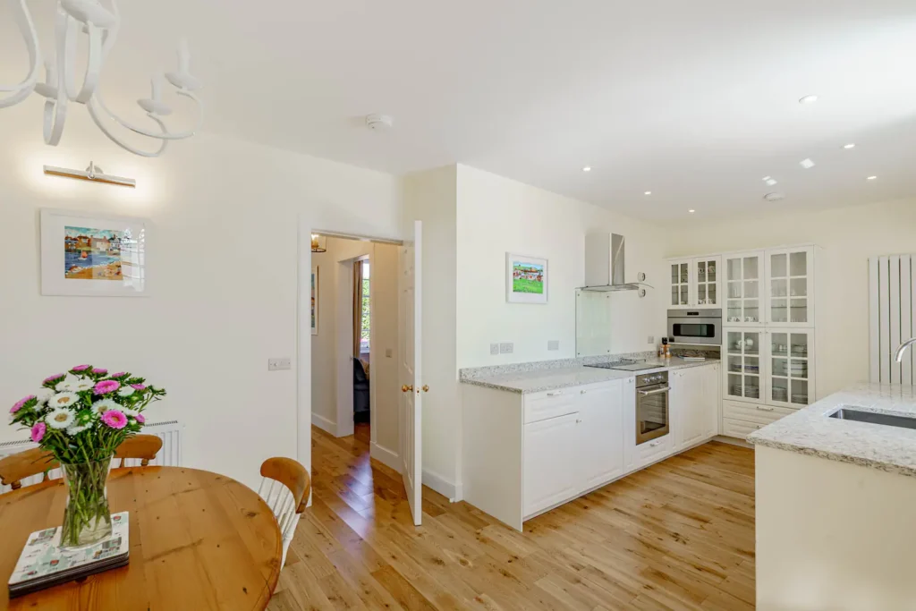 Bright modern kitchen and dining area with wooden flooring, white cabinets, and a round table with flowers.