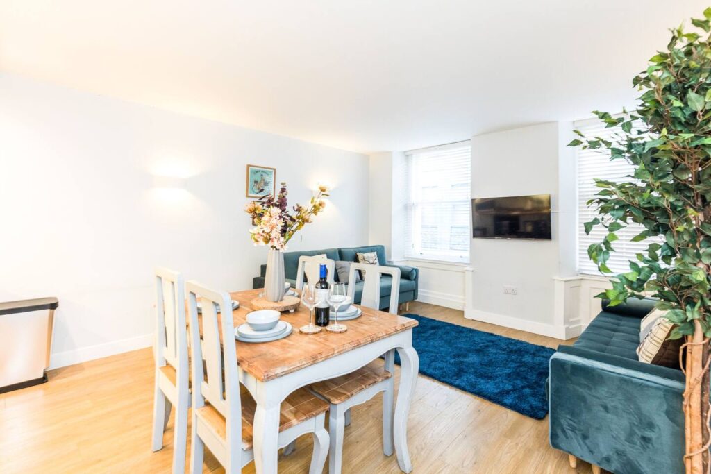 Modern dining and living area with wooden table, blue sofas, and a wall-mounted TV in a bright, stylish apartment.