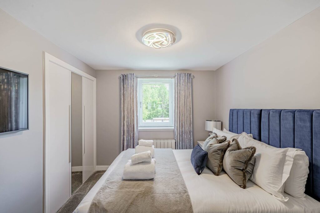 Modern bedroom with navy headboard, stylish light fixture, and window view, featuring plush pillows and folded towels.