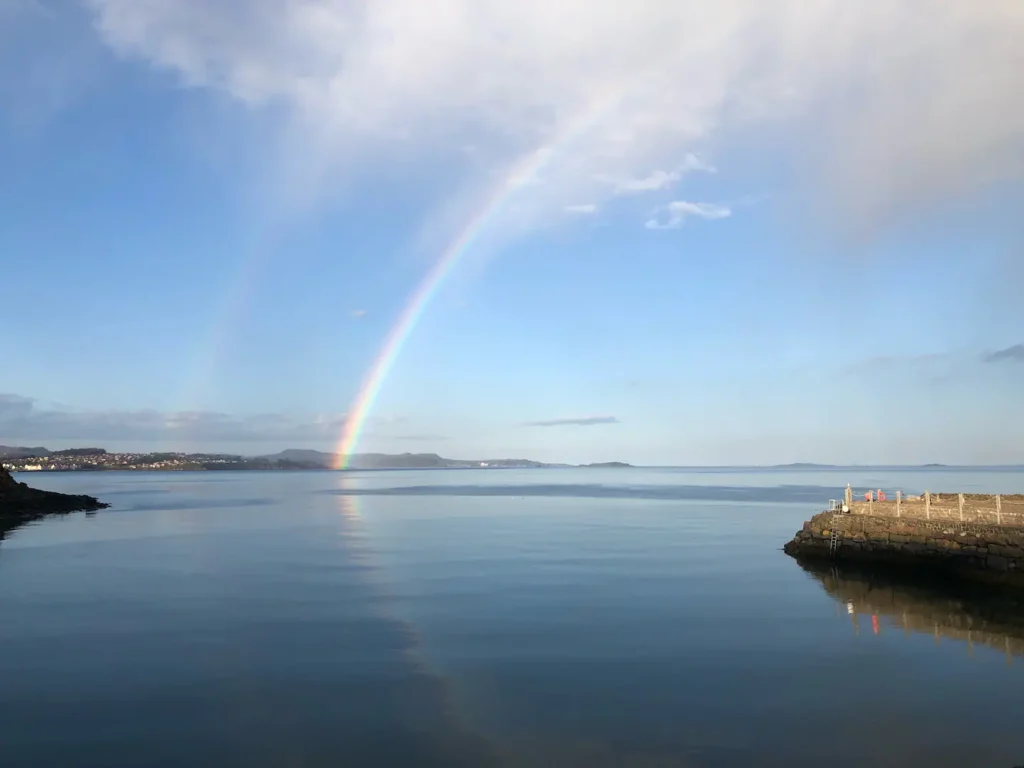 Colorful rainbow over calm sea with distant coastline and stone pier under a clear blue sky.