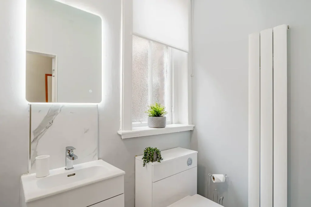 Modern bathroom with illuminated mirror, sleek vanity, and potted plants on the window sill, enhancing elegance.