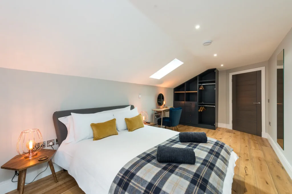 Cozy modern loft bedroom with double bed, plaid blanket, and skylight. Elegant workspace and open closet in view.