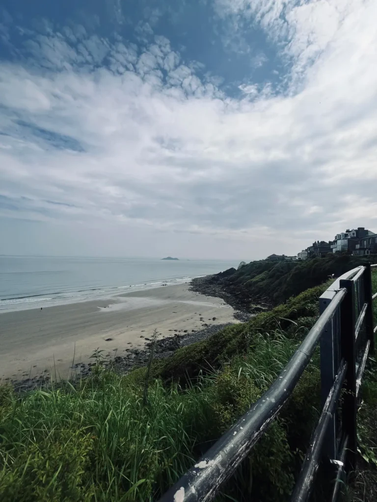 Coastal landscape with cloudy sky, sandy beach, green cliffs, and a distant island in view, taken from a metal railing viewpoint.