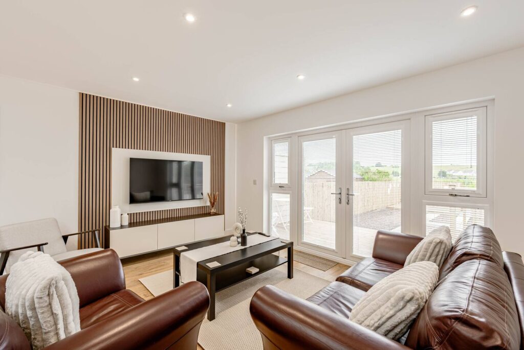 Modern living room with leather sofas, wall-mounted TV, and large windows offering natural light.