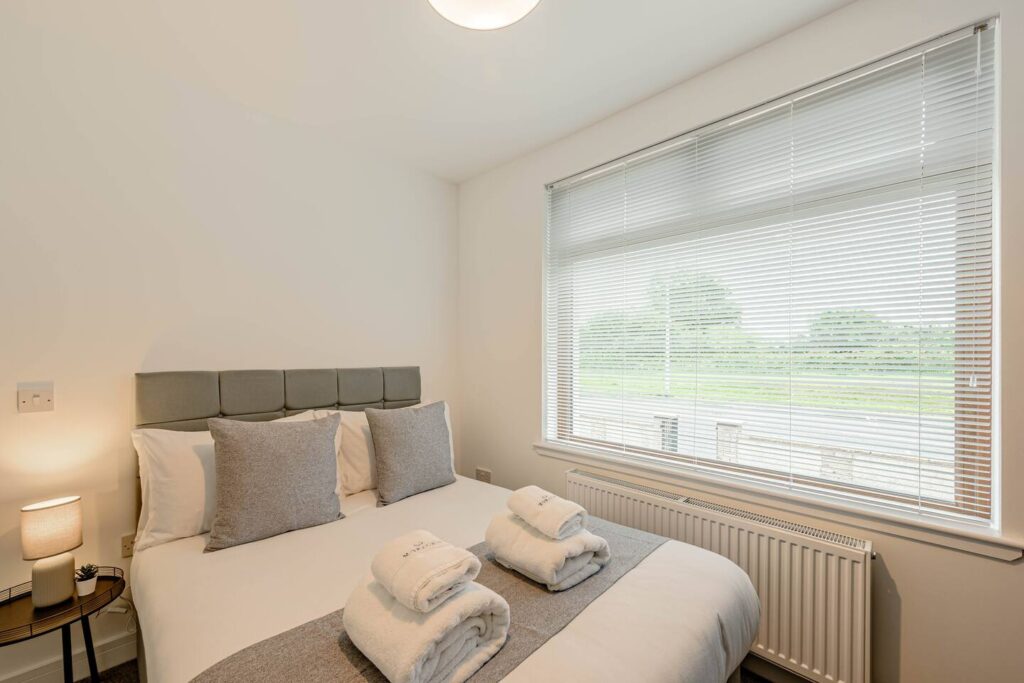 Cozy bedroom with a modern bed, fluffy towels, and a large window with blinds overlooking a green landscape.