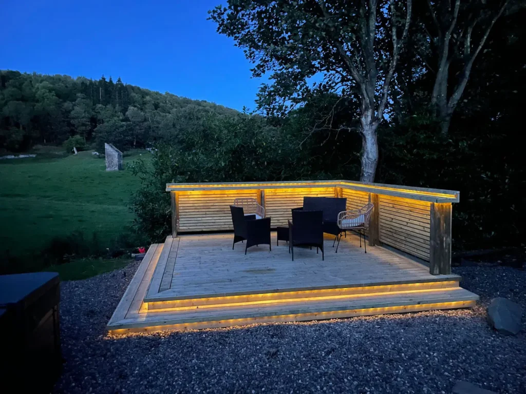 Cozy outdoor deck with warm lighting and seating, set against a scenic evening backdrop of trees and hills.