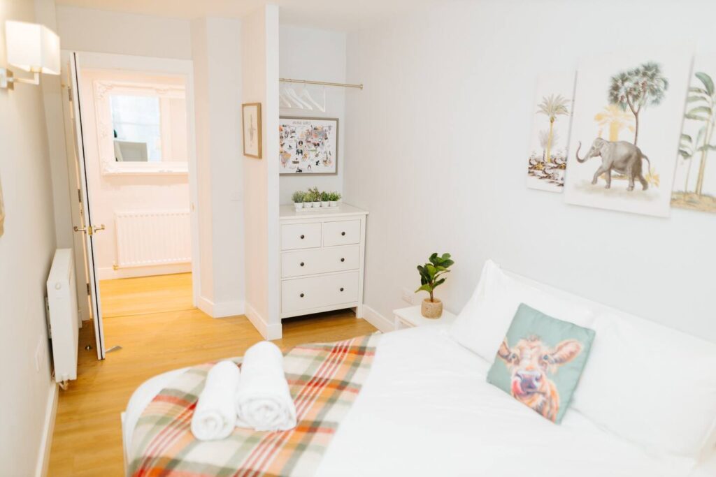 Bright, cozy bedroom with white decor, plants, and animal-themed art on walls. Pillow and towels on bed.