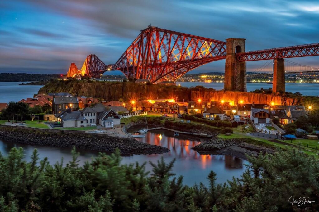 Illuminated red bridge over water at dusk, with a village in the foreground, reflecting a vibrant evening sky.