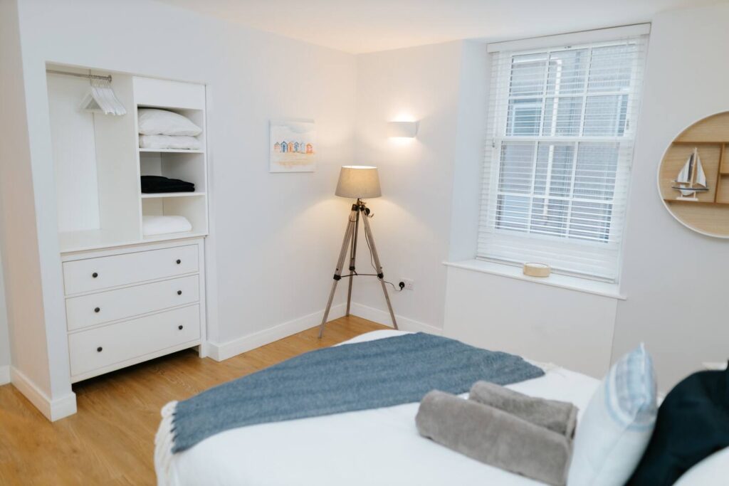 Modern bedroom with white furnishings, wooden floors, and natural light from a window. Cozy and minimal design.