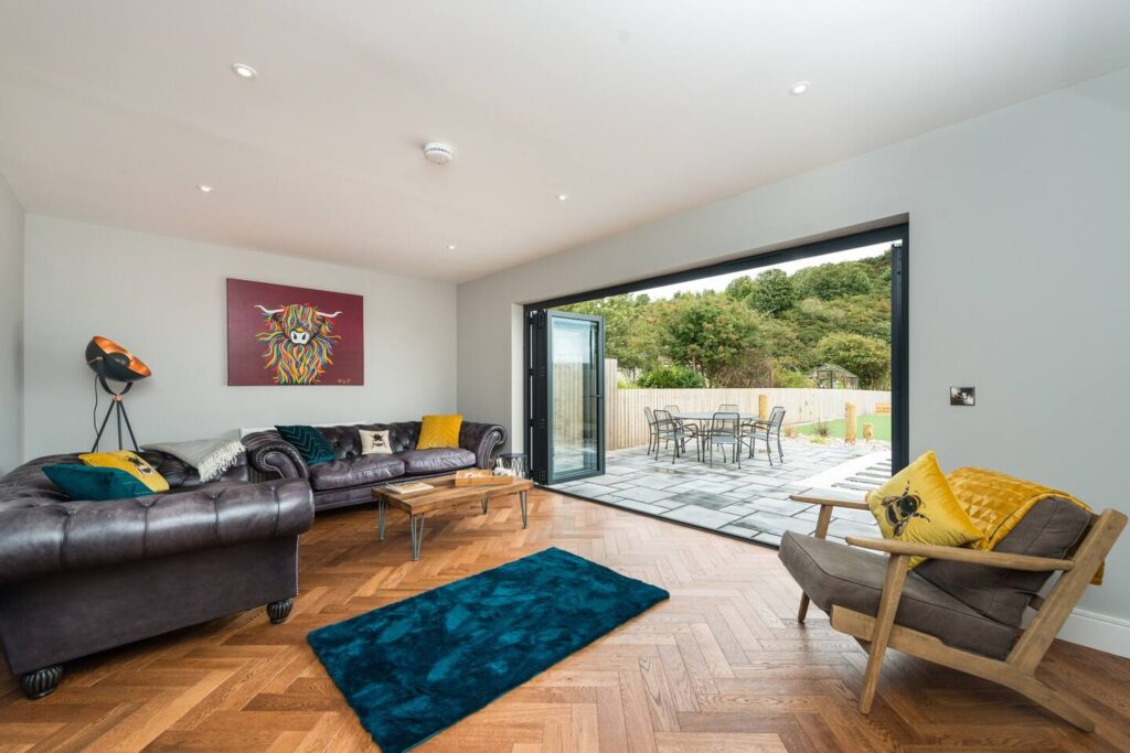 Modern living room with open patio doors, brown leather sofas, and colorful artwork on white walls.