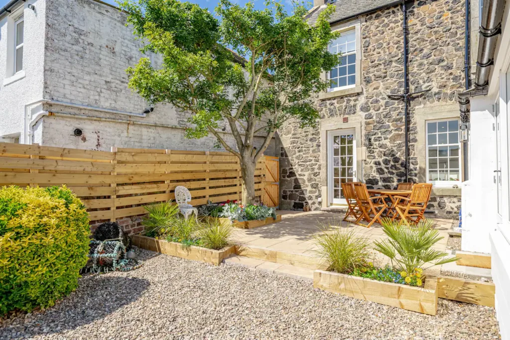 Cozy stone patio with wooden furniture, garden plants, and a tree, enhancing a charming outdoor setting.