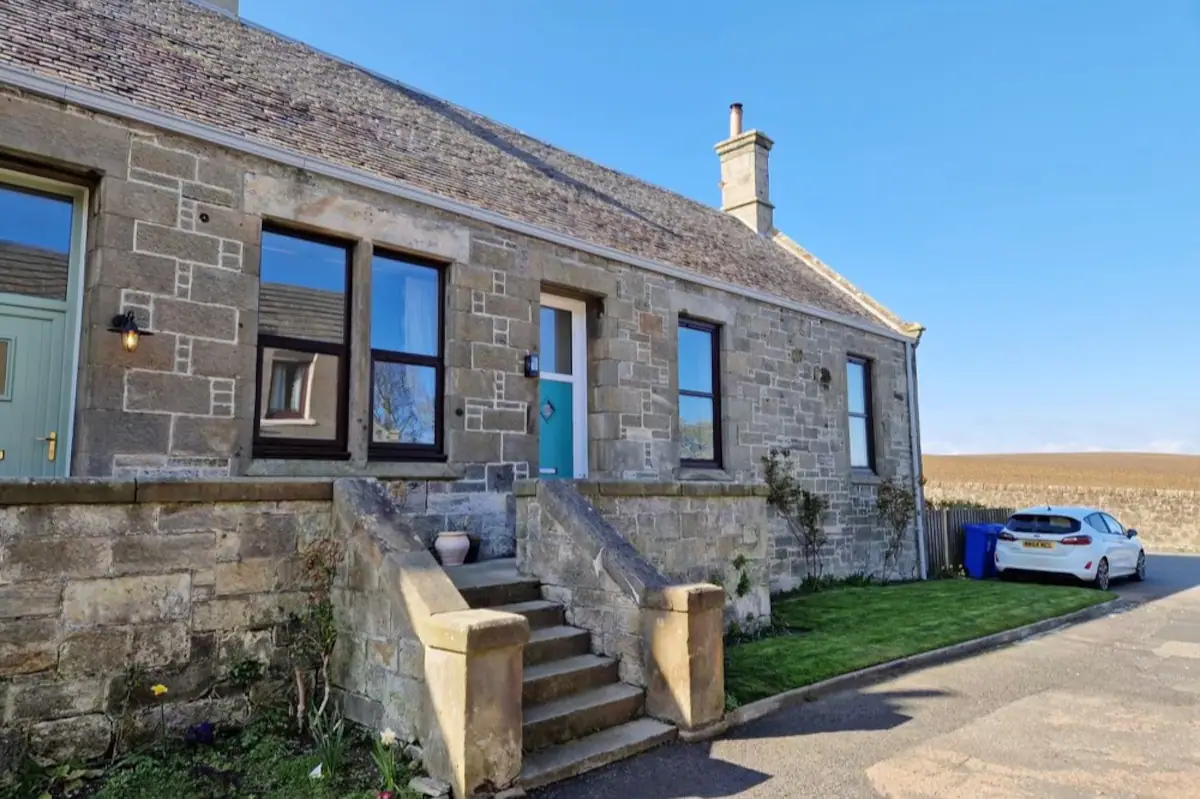 Stone cottage with blue door, front steps, and parked car on a sunny day.