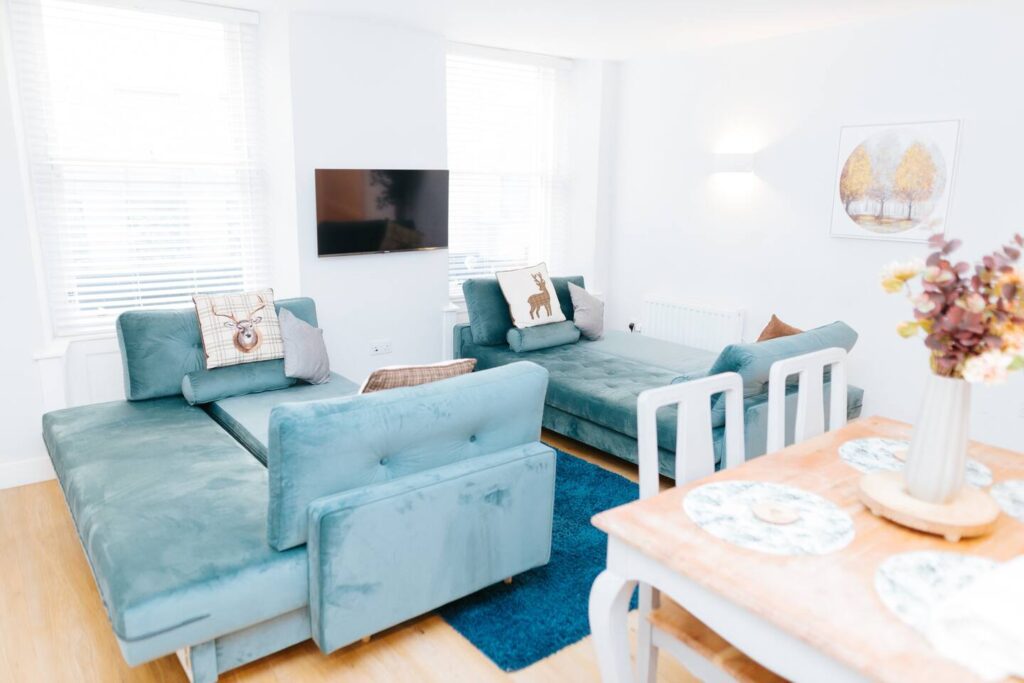 Bright living room with teal sofas, wall art, TV, and dining area featuring floral arrangement. Cozy modern interior design.