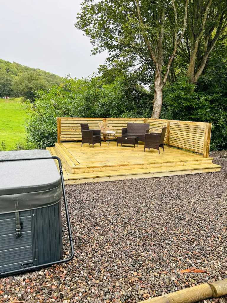 Wooden deck with outdoor seating, surrounded by green trees, next to a hot tub on pebble ground.