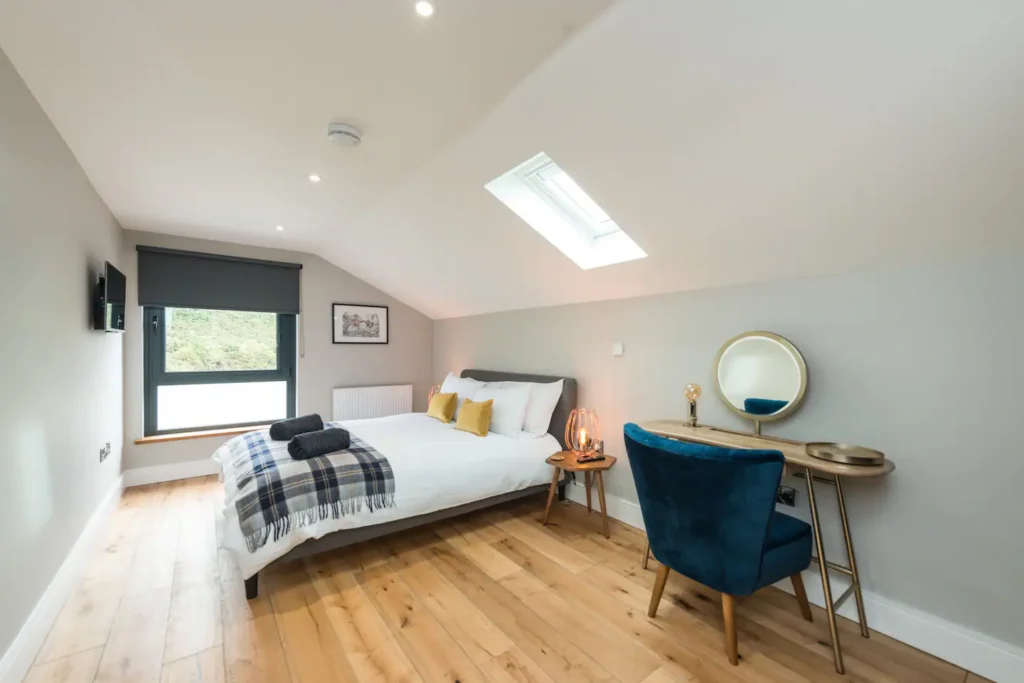 Modern bedroom with skylight, cozy bed, teal chair, and wooden desk. Bright and inviting space with natural light.