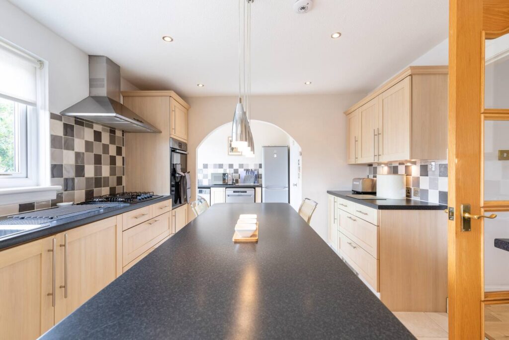 Modern kitchen with wooden cabinets, black countertops, and stainless steel appliances under pendant lighting.