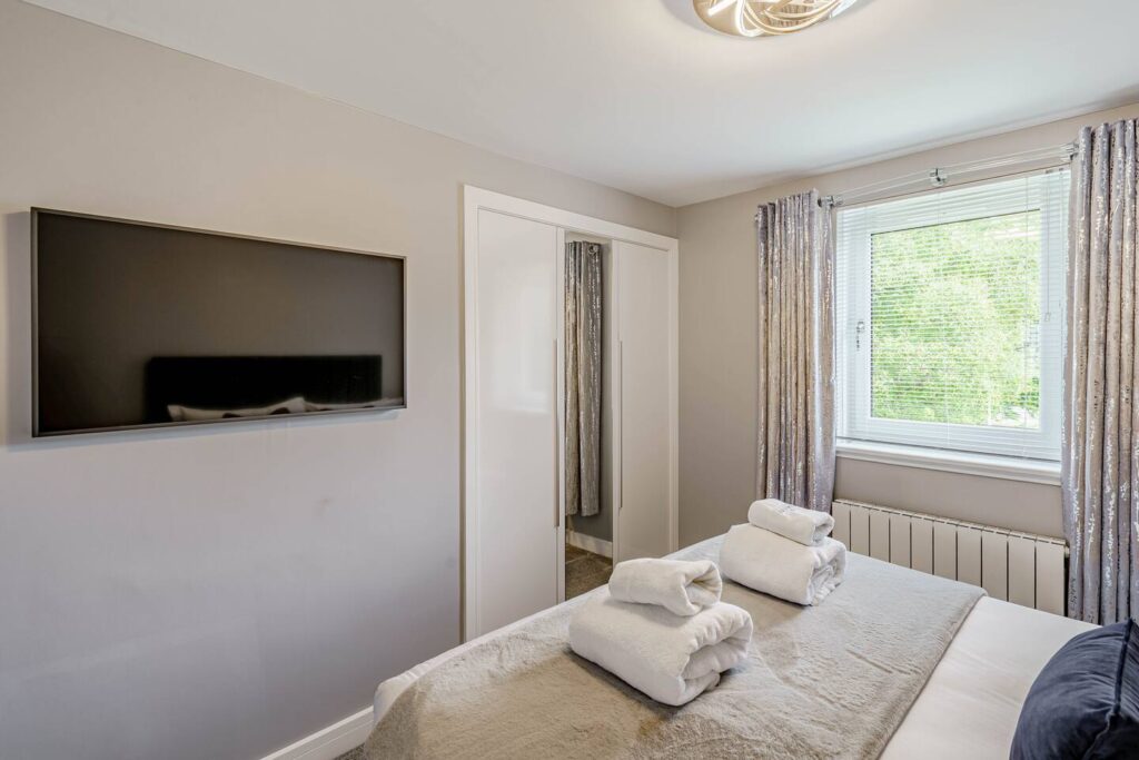 Modern bedroom with neatly folded towels on a bed, wall-mounted TV, and natural light through window.