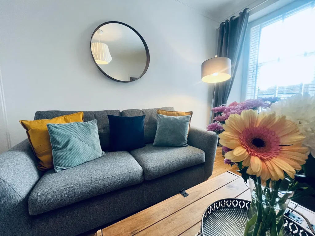 Stylish living room with cozy grey sofa, colorful cushions, round mirror, and fresh flowers on a wooden table.