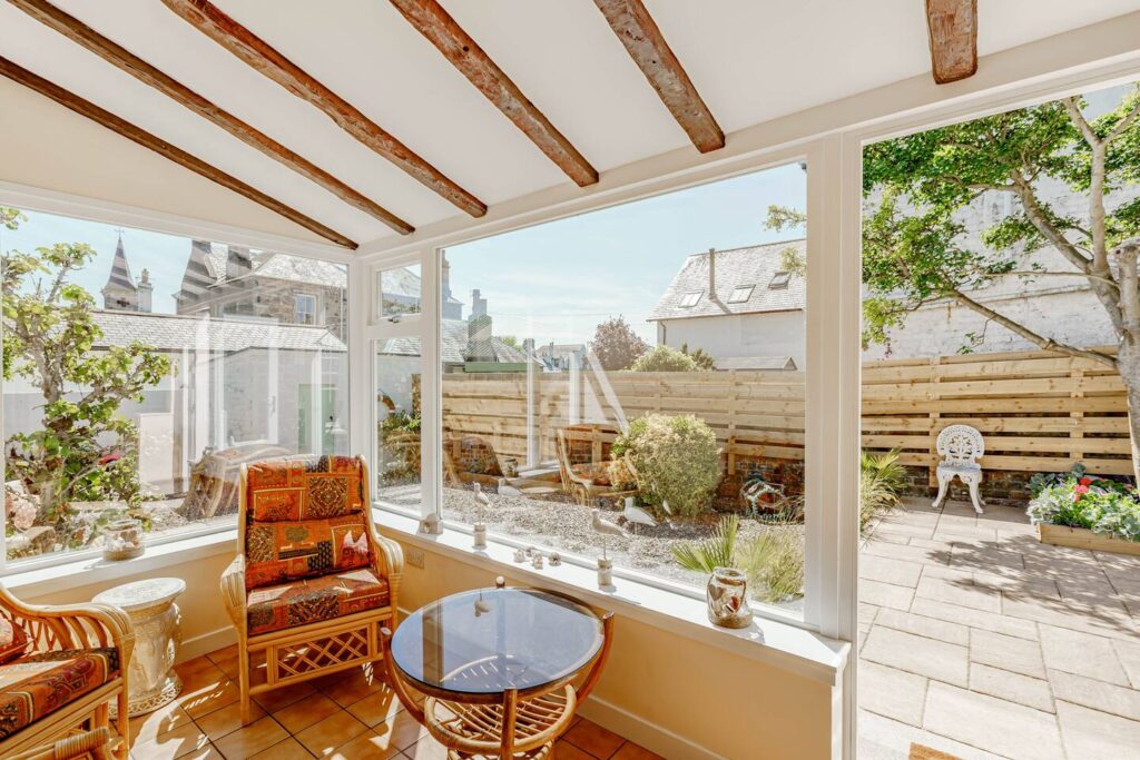 Cozy indoor sunroom with rattan seating and garden view, featuring rustic wooden beams and large windows.