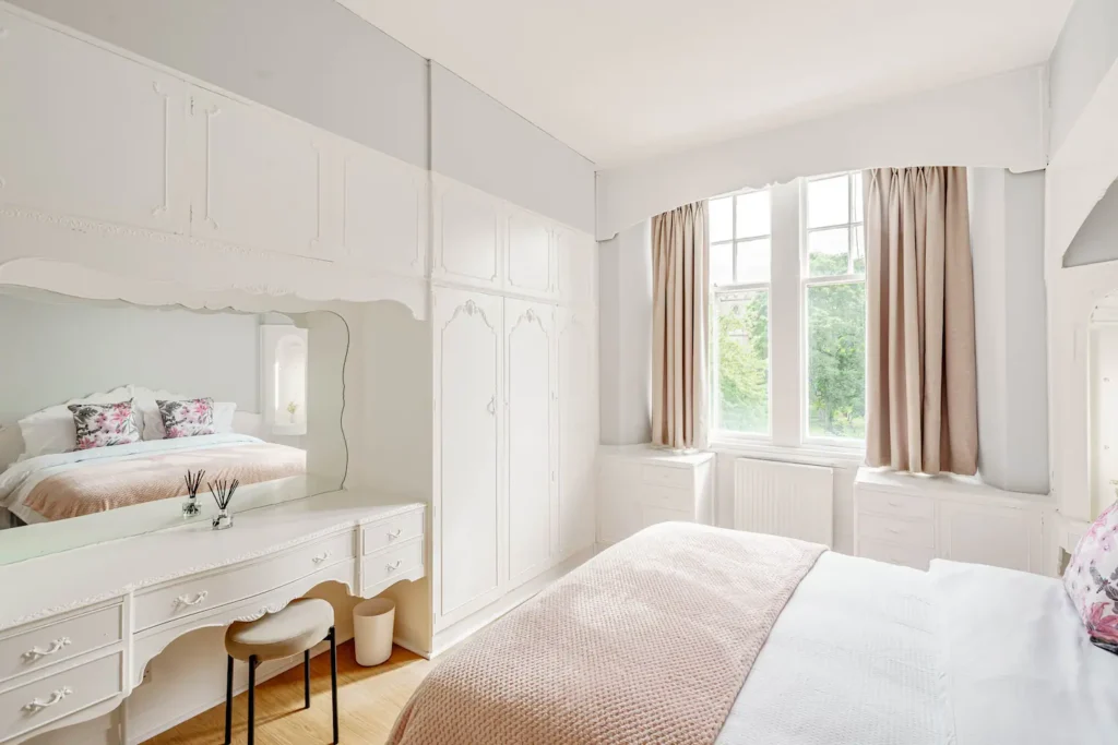 Bright bedroom with white decor, large windows, and a cozy bed with floral pillows. Elegant vanity with mirror.