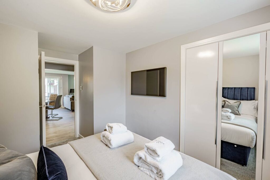 Modern bedroom with towels on bed, wall-mounted TV, and mirrored closet reflecting a cozy bed setup.