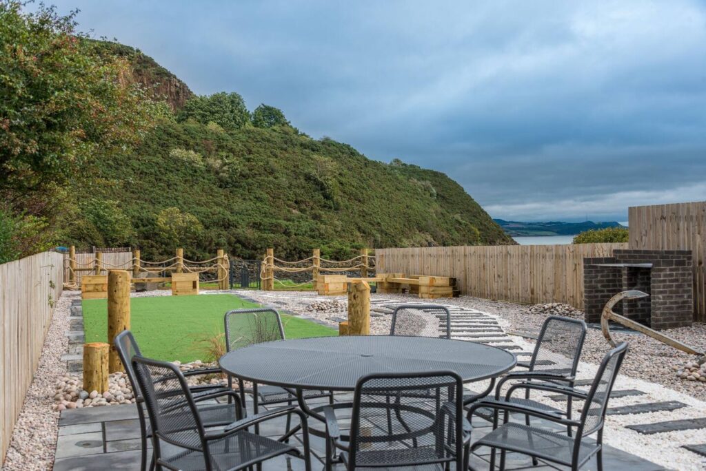 Outdoor patio with round table, chairs, and mountain view, surrounded by greenery and wooden fences under cloudy skies.