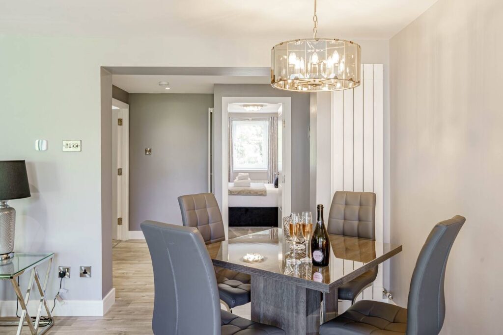 Modern dining area with sleek gray chairs, glass table, and pendant light. Champagne and glasses add elegance.