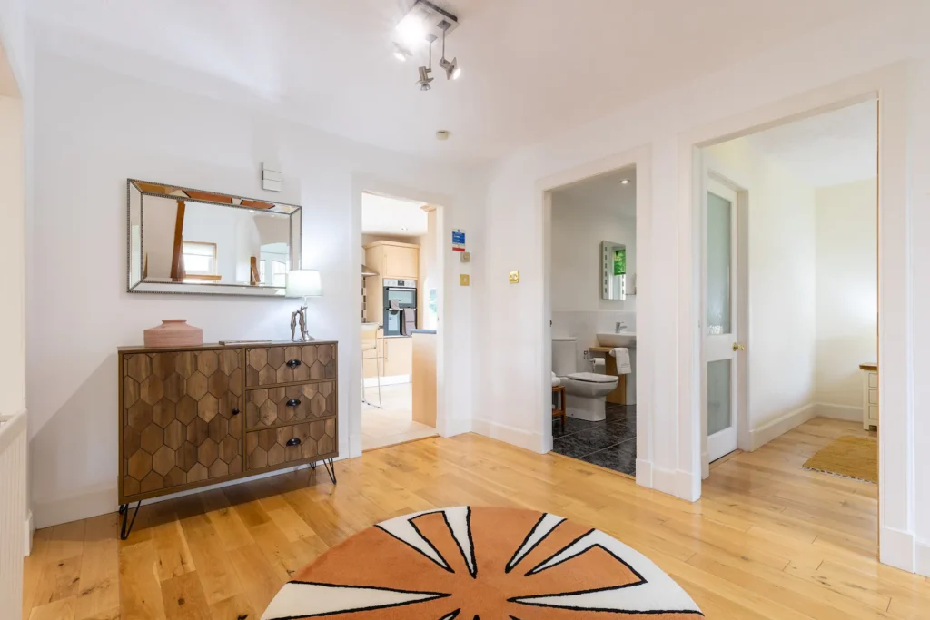 Modern hallway interior with wood flooring, stylish dresser, round rug, mirror, leading to kitchen and bathroom.