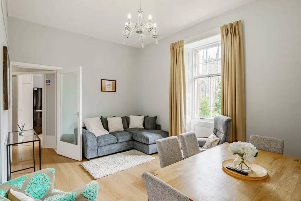 Bright living room with modern gray sofa, wooden dining table, chandelier, and large window with beige curtains.