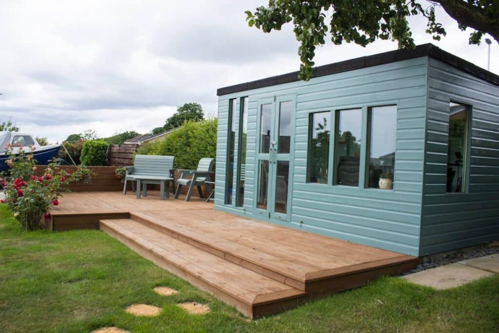 Modern garden office with wood deck and outdoor seating surrounded by lush greenery and flowers.