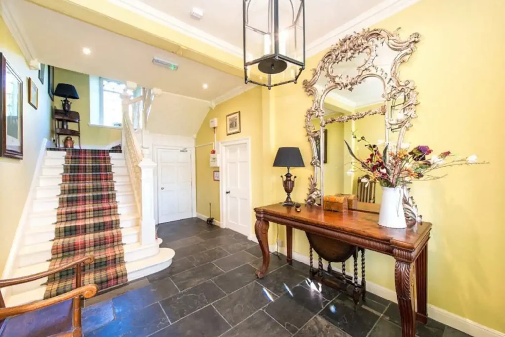 Elegant hallway with vintage mirror, wooden console, plaid staircase, and yellow walls creating a warm, classic ambiance.