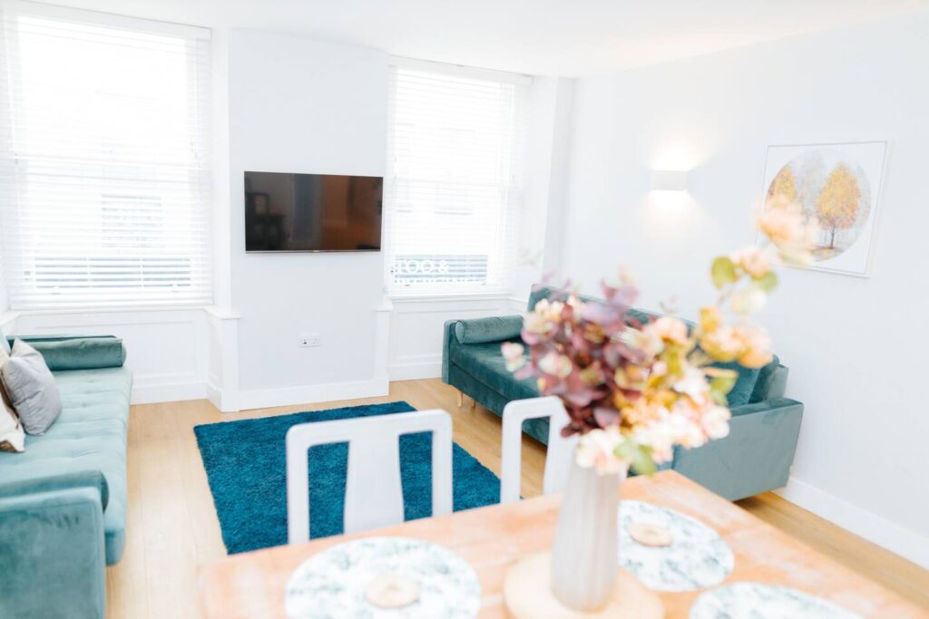 Bright living room with teal sofas, wall-mounted TV, and vibrant floral centerpiece on dining table.