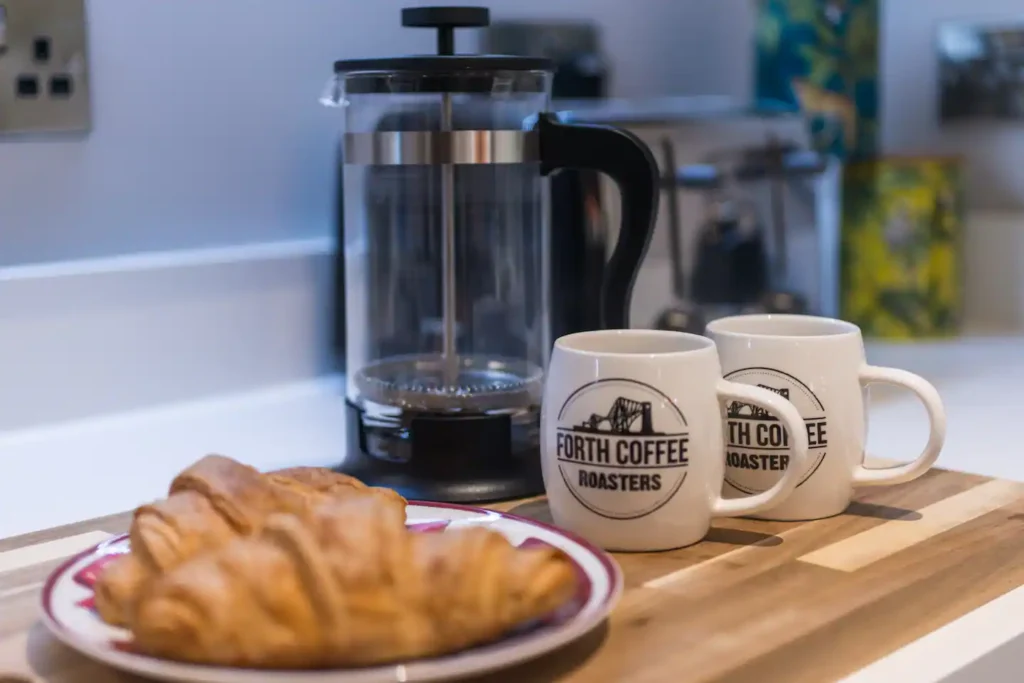 French press with two mugs from Forth Coffee Roasters and croissants on a wooden board in a cozy kitchen setting.