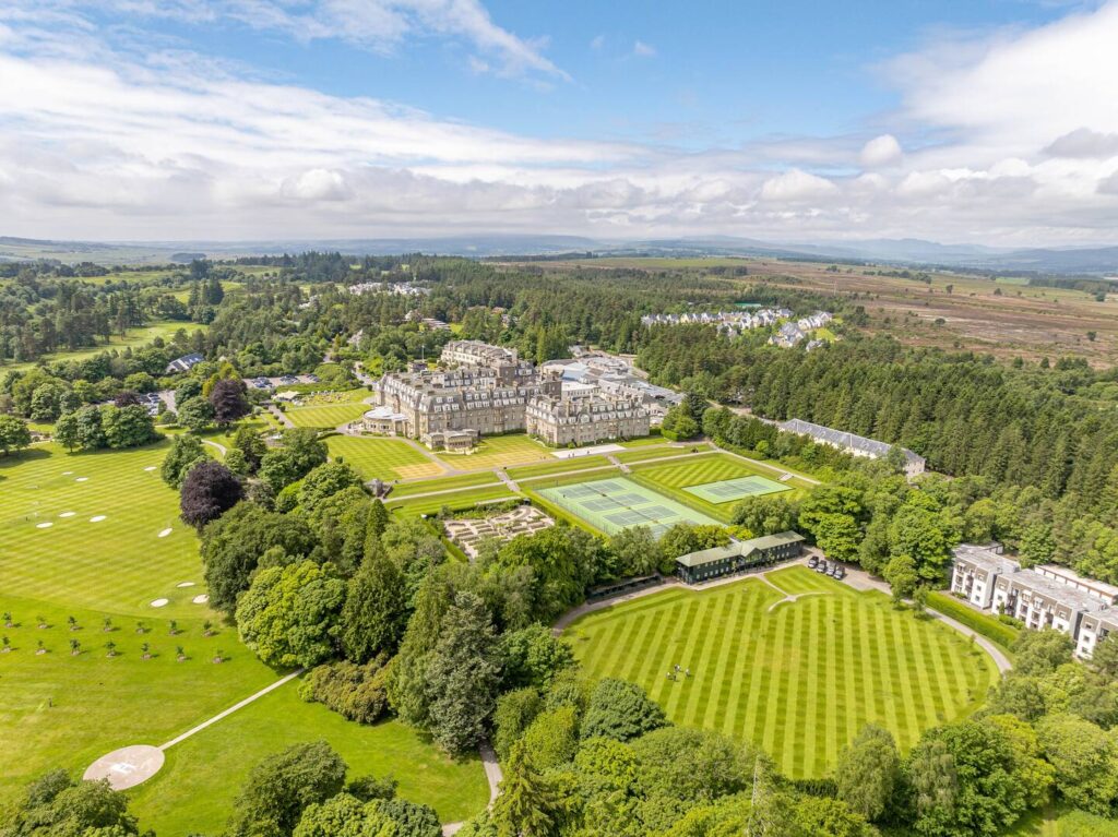 Aerial view of a luxury hotel with landscaped gardens, tennis courts, and surrounding lush greenery on a sunny day.