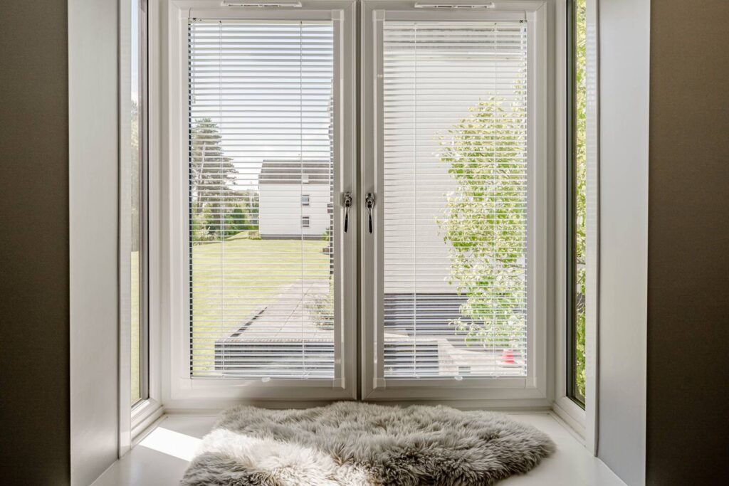 Sunny window view with blinds, fluffy rug on sill, overlooking a lawn and distant building. Serene indoor ambiance.