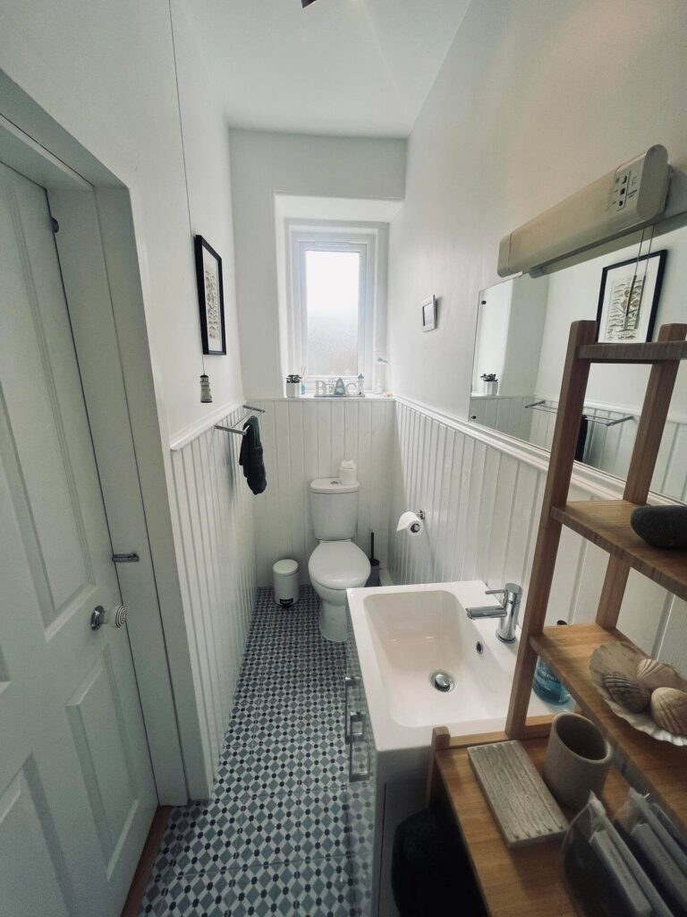 Compact bathroom with white paneling, patterned floor tiles, a window, small sink, and shelving with decorative items.