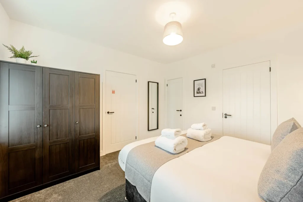 Modern bedroom with dark wardrobe, neatly made bed, white decor, and mirror for minimalist style.