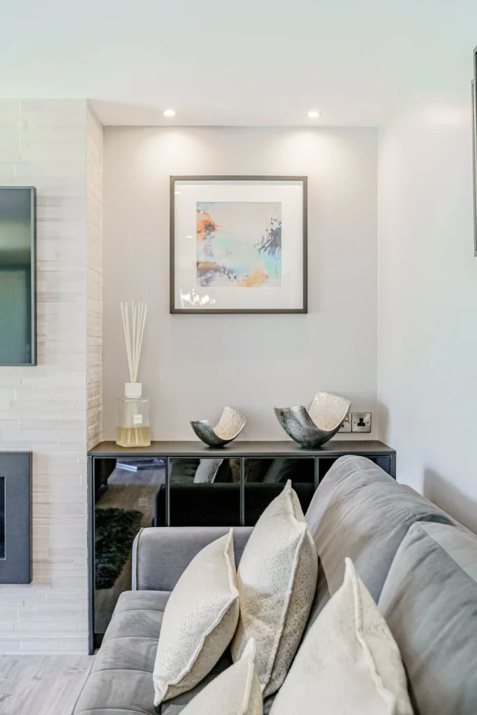 Modern living room with grey sofa, decorative cushions, abstract art, and stylish black cabinet under soft lighting.