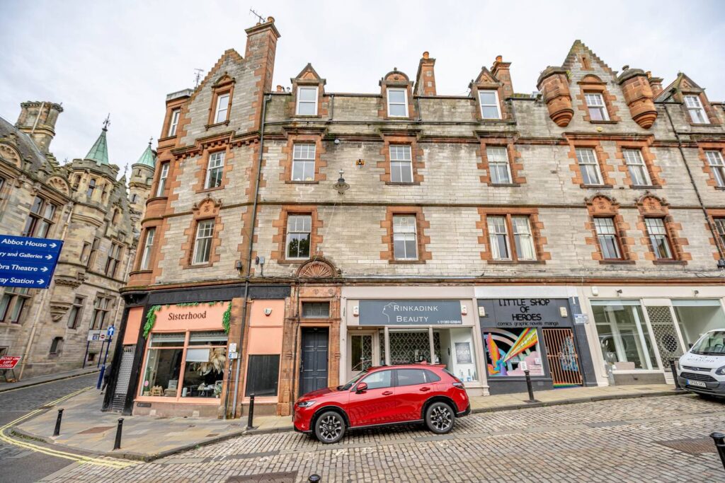 Historic city street with shops and a parked red car, featuring vintage architecture and cobblestone pavement.