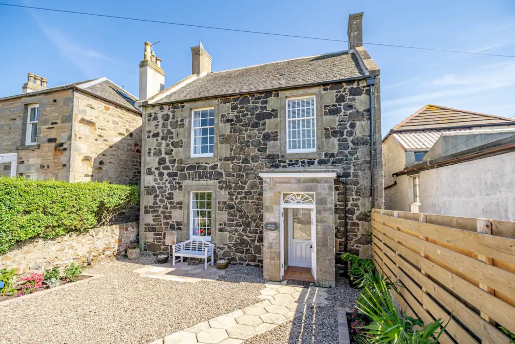 Charming stone cottage with garden and seating area under a sunny blue sky, perfect for a relaxing getaway.