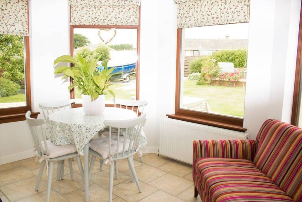 Bright sunroom with table and chairs, plants, and striped couch overlooking garden through large windows.
