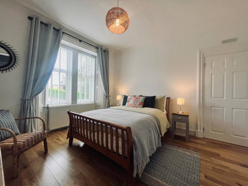 Cozy bedroom with wooden bed, soft lighting, wicker chair, and large window with curtains.