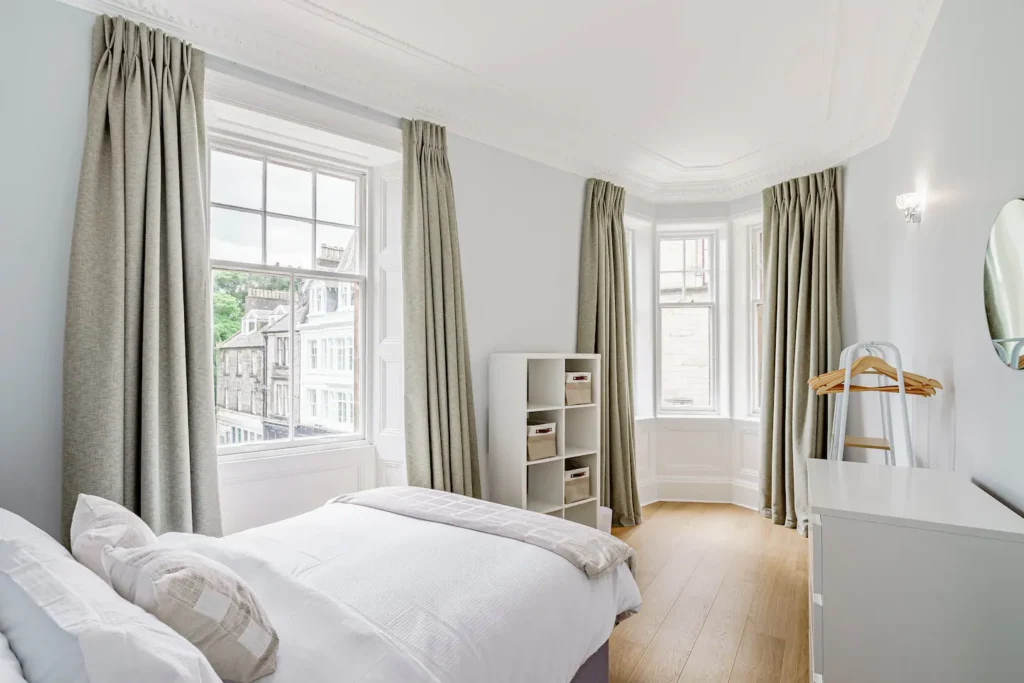 Bright, minimalist bedroom with large windows, white walls, wood floor, and neutral tones. Cozy and inviting atmosphere.