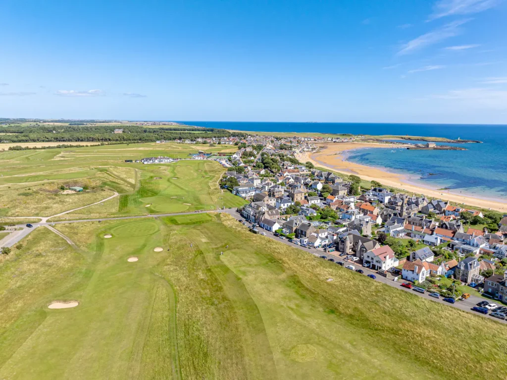 Coastal town with golf course and sandy beach under clear blue sky, aerial view.