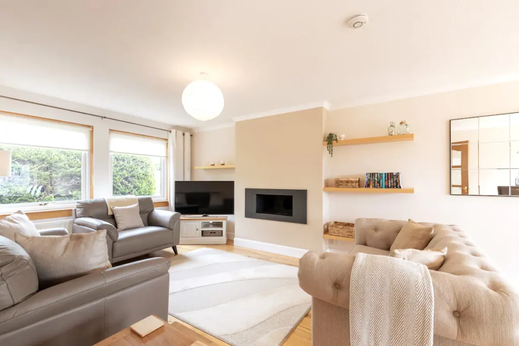 Cozy modern living room with beige sofas, fireplace, TV, and natural light through large windows.