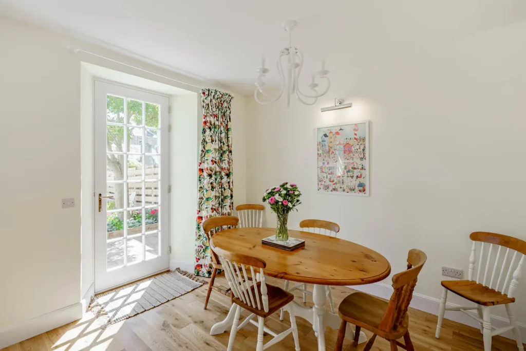 Bright dining area with wooden table, chairs, floral curtains, and French doors leading to a garden.