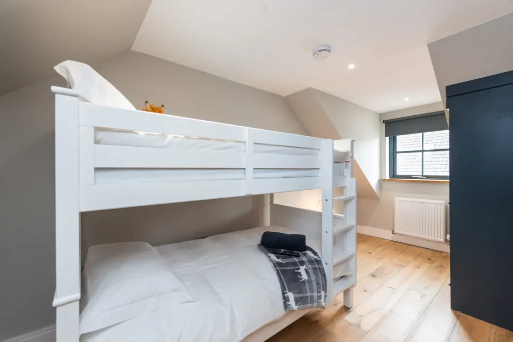 Cozy attic bedroom with white bunk beds, wooden floor, and natural light from the window.