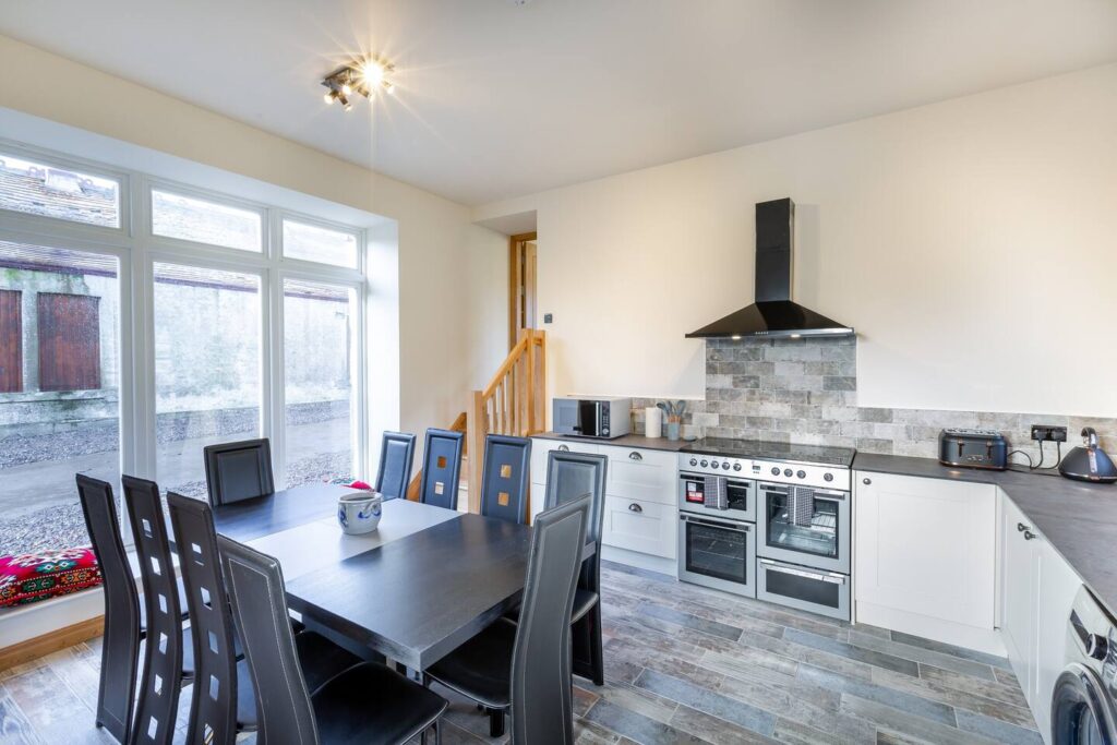 Modern kitchen with dining table, black chairs, large windows, and stainless steel appliances under a tiled backsplash.