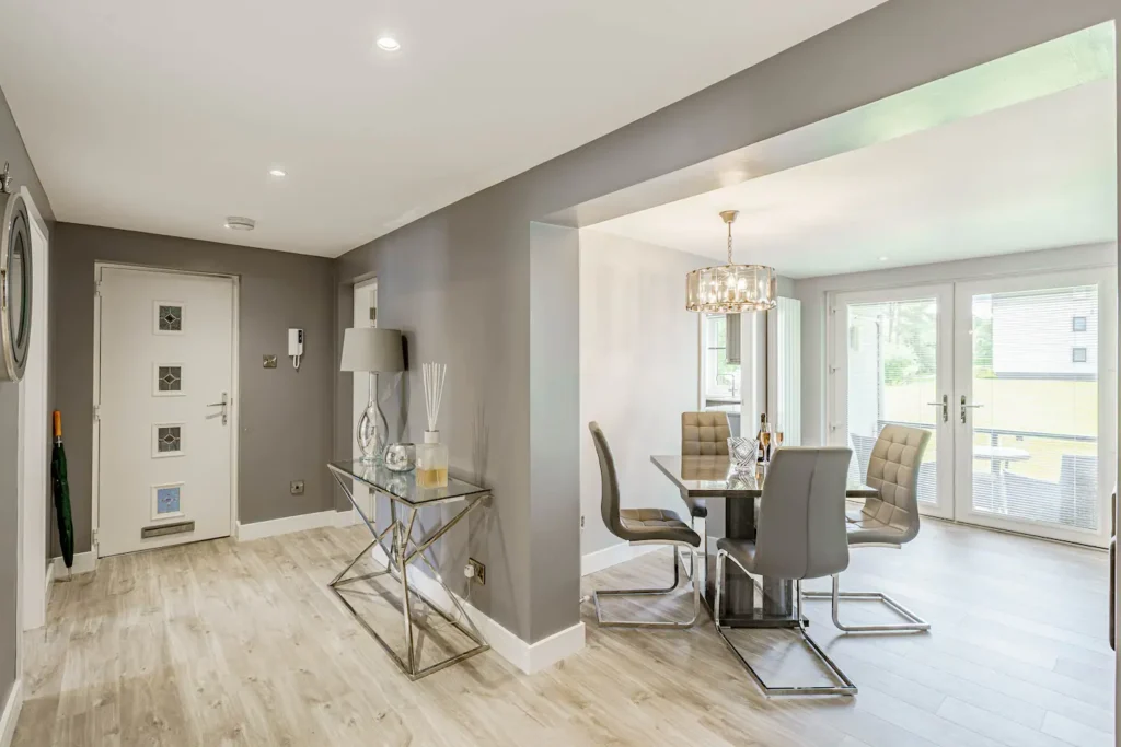 Modern dining room with glass table, gray chairs, and chandelier lighting, adjacent to a sleek entrance with decor.